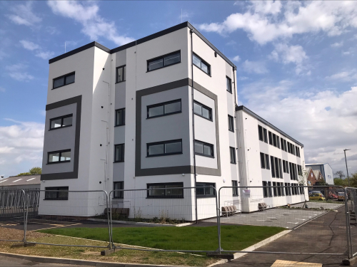 Four storey modern white apartment block with grass and concrete area outside.