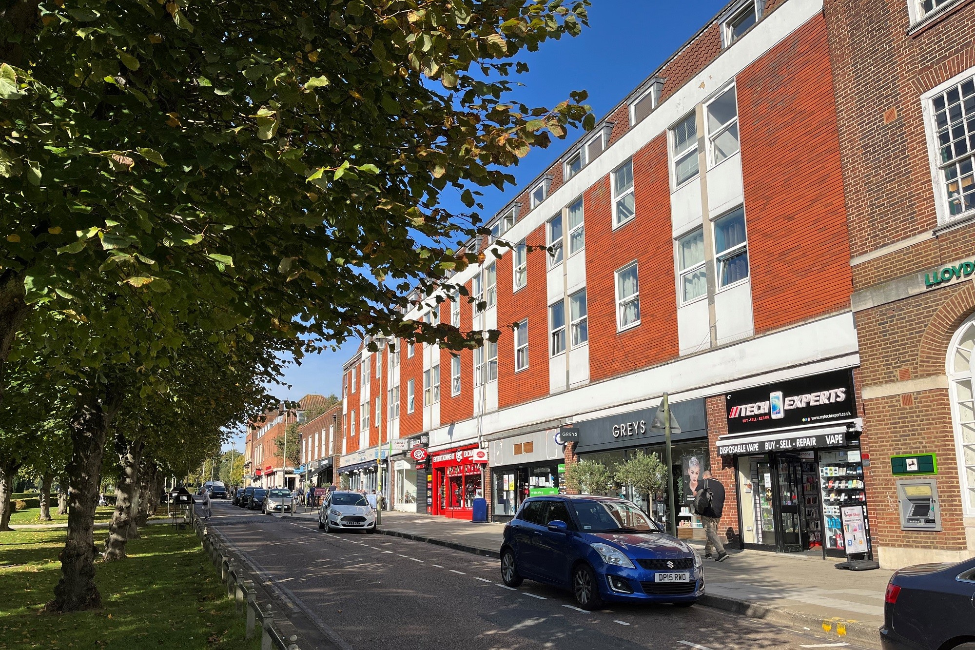 A street in wgc town centre where you can see cars parked on the side of the road and shops