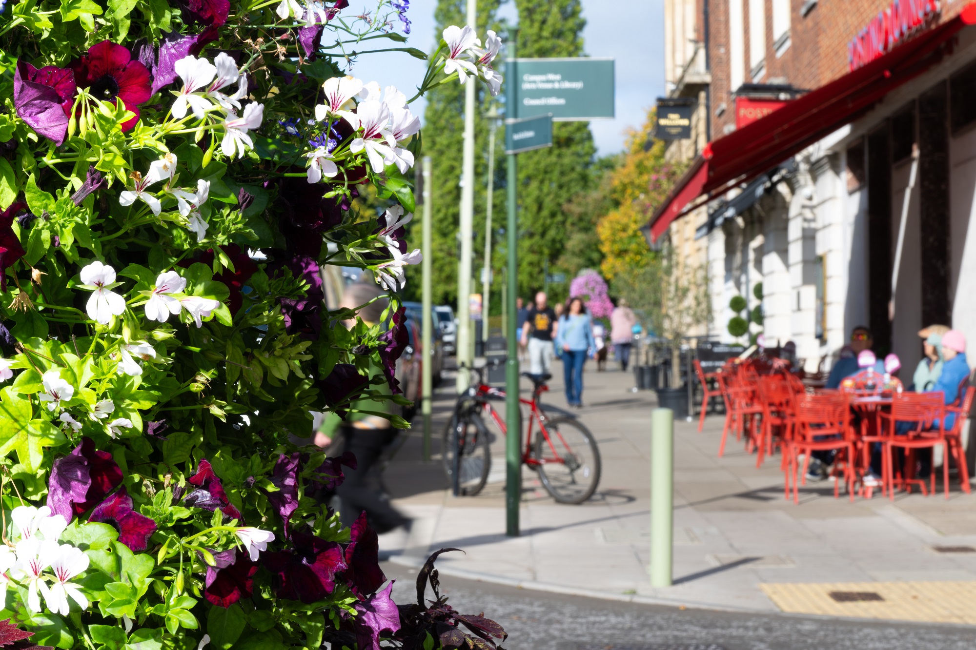 Street in WGC town centre. Purple and white folowers on the left side with people sitting on red chairs on the left side.
